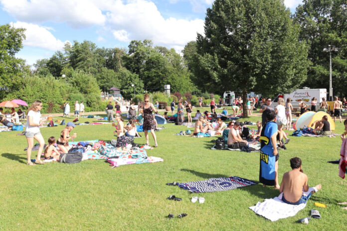 Viele verschiedene Badegäste befinden sich gemeinsam auf einer Liegewiese, einige sind mit Badebekleidung bekleidet, die Liegewiese befindet sich in einem Freibad.