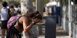 Eine braungebrannte junge attraktive Frau kühlt sich bei höllischen Temperaturen in der Innenstadt mit dem Wasser eines Brunnens ab. Sie trägt wenig Kleidung und einen Sportbeutel auf dem Rücken. Es herrscht offensichtlich sommerliche Hitze.