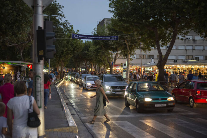 Mehrere Fußgänger gehen über eine mehrspurige Straße über die Ampel. Es dämmert schon und die Autos haben ihre Scheinwerfer an. Im Hintergrund sieht man einen beleuchteten Markt. Eine neue Bußgeldwelle kommt nun auf Millionen Autofahrer zu.