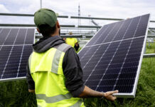 Ein Arbeiter mit Kappe und gelber Sicherheitsweste transportiert und befestigt auf einer Baustelle auf einer Wiese ein Solarpanel für eine größere Solaranlage.