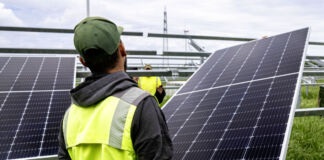 Ein Arbeiter mit Kappe und gelber Sicherheitsweste transportiert und befestigt auf einer Baustelle auf einer Wiese ein Solarpanel für eine größere Solaranlage.