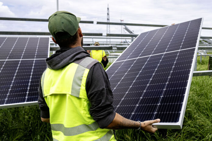 Ein Arbeiter mit Kappe und gelber Sicherheitsweste transportiert und befestigt auf einer Baustelle auf einer Wiese ein Solarpanel für eine größere Solaranlage.