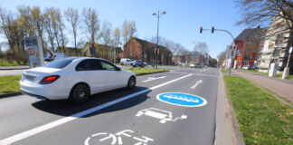 Ein weißes Auto fährt auf einer zweispurigen Straße in der Nähe eines Dorfes. Auf der Spur rechts vom Auto sind mehrere Symbole mit Farbe angezeichnet. Ein Fahrrad, eine E-Ladestation und ein Bus-Kennzeichen.