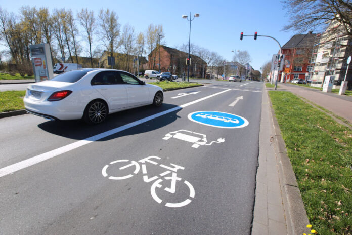 Ein weißes Auto fährt auf einer zweispurigen Straße in der Nähe eines Dorfes. Auf der Spur rechts vom Auto sind mehrere Symbole mit Farbe angezeichnet. Ein Fahrrad, eine E-Ladestation und ein Bus-Kennzeichen.