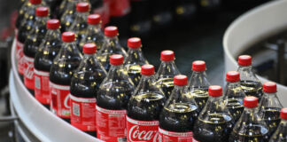 Der Blick in eine Fabrik. Über das Fabrikband fahren mehrere Flaschen von originaler Coca-Cola. Sie sind frisch gefüllt und offenbar für den Abtransport bereit. Die Flaschen der Cola sind aus Plastik und in dem typischen Coca-Cola Rot-Weiß gehalten.