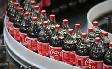 Der Blick in eine Fabrik. Über das Fabrikband fahren mehrere Flaschen von originaler Coca-Cola. Sie sind frisch gefüllt und offenbar für den Abtransport bereit. Die Flaschen der Cola sind aus Plastik und in dem typischen Coca-Cola Rot-Weiß gehalten.