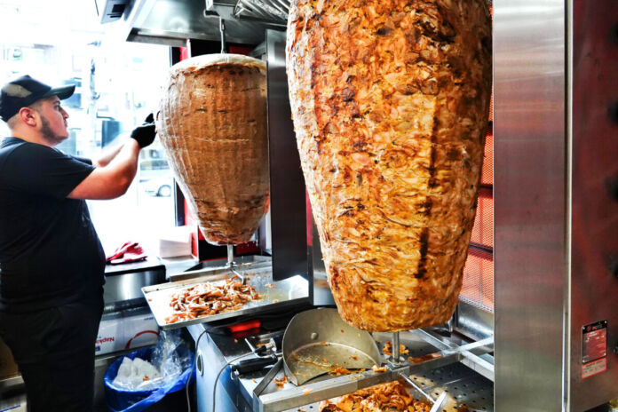 In einem Dönerladen drehen sich saftige Dönerspieße, während ein Verkäufer und Mitarbeiter Fleisch vom Dönerspieß abschneidet um es in einem leckeren Döner zu servieren und an seine Kunden auf der Straße zu verkaufen.