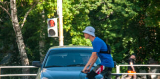 Ein Fahrradfahrer mit Hut und Brusttasche überquert im Sommer auf seinem Drahtesel eine Straße. Ein Auto wartet geduldig auf sein Passieren, während im Hintergrund ein Wald und eine rote Ampel zu sehen ist.