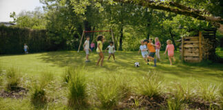 Zahlreiche Kinder spielen ausgelassen auf einer grünen Wiese neben einer Schaukel Fußball. Es sind Mädchen und Jungen dabei und der Ball rollt beim herrlichen Wetter und unter Jubel der Kleinen.