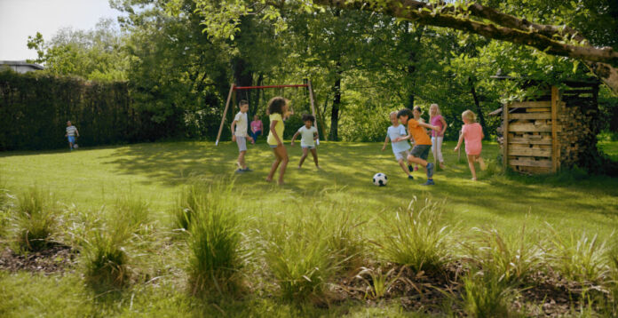 Zahlreiche Kinder spielen ausgelassen auf einer grünen Wiese neben einer Schaukel Fußball. Es sind Mädchen und Jungen dabei und der Ball rollt beim herrlichen Wetter und unter Jubel der Kleinen.