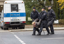 Eine Gruppe Polizisten führt einen Demonstranten oder Aktivisten und wahrscheinlichen Klimakleber von der Straße. Im Hintergrund ist ein Polizeifahrzeug zu sehen. Der abgeführte Mann wird getragen und hat ein verkrampftes Gesicht.