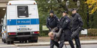 Eine Gruppe Polizisten führt einen Demonstranten oder Aktivisten und wahrscheinlichen Klimakleber von der Straße. Im Hintergrund ist ein Polizeifahrzeug zu sehen. Der abgeführte Mann wird getragen und hat ein verkrampftes Gesicht.