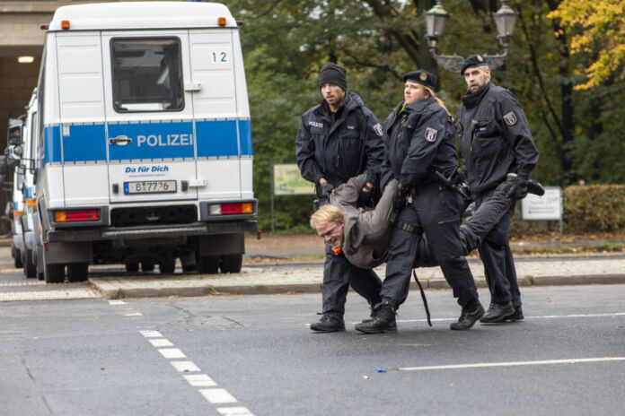 Eine Gruppe Polizisten führt einen Demonstranten oder Aktivisten und wahrscheinlichen Klimakleber von der Straße. Im Hintergrund ist ein Polizeifahrzeug zu sehen. Der abgeführte Mann wird getragen und hat ein verkrampftes Gesicht.