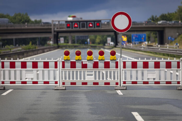 Auf einer leeren Autobahn ist eine rot-weiße Baustellenabsperrung zu sehen, die die Straße für den Verkehr sperrt. Ein Haltestoppschild weist extra darauf hin, dass hier nicht gefahren wird.