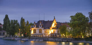 In einer abendlichen Atmosphäre steht an einem See ein schönes Gebäude, ein Wirtshaus mit Außenbestuhlung. Es ist ein Wirtshaus samt Biergarten, das abends zahlreiche Gäste und Trinklustige anlockt.