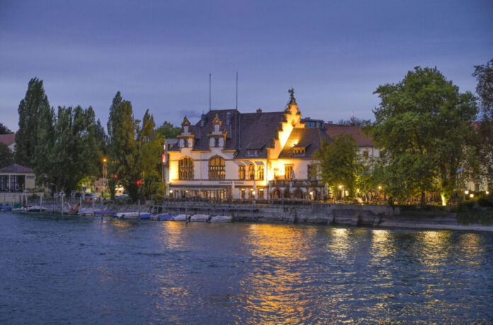 In einer abendlichen Atmosphäre steht an einem See ein schönes Gebäude, ein Wirtshaus mit Außenbestuhlung. Es ist ein Wirtshaus samt Biergarten, das abends zahlreiche Gäste und Trinklustige anlockt.