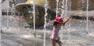 Ein kleines Mädchen mit einem pinkfarbenen Sonnenhut gönnt sich eine Abkühlung. Es steht in einem Springbrunnen und läuft durch das Wasser. Rechts und links spritzen Wasserfontänen das Kind an und kühlen es so ab.