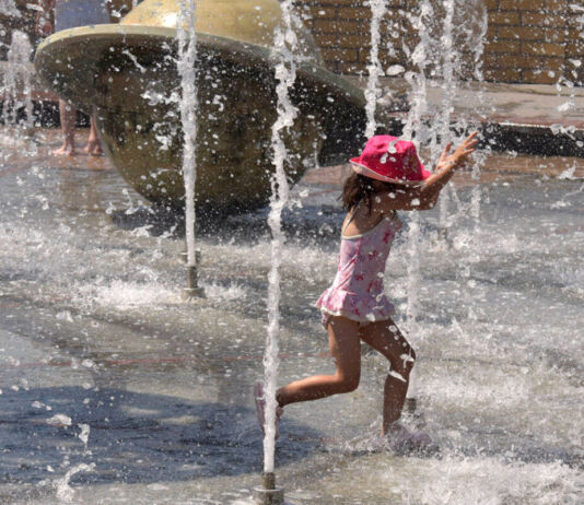 Ein kleines Mädchen mit einem pinkfarbenen Sonnenhut gönnt sich eine Abkühlung. Es steht in einem Springbrunnen und läuft durch das Wasser. Rechts und links spritzen Wasserfontänen das Kind an und kühlen es so ab.