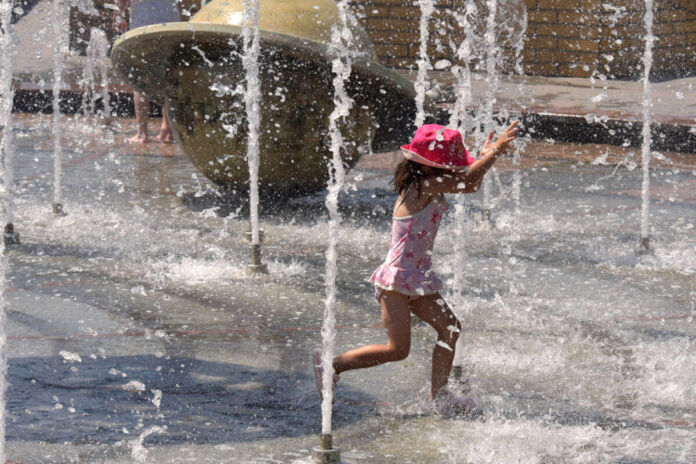 Ein kleines Mädchen mit einem pinkfarbenen Sonnenhut gönnt sich eine Abkühlung. Es steht in einem Springbrunnen und läuft durch das Wasser. Rechts und links spritzen Wasserfontänen das Kind an und kühlen es so ab.