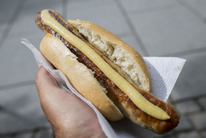 Eine Hand hält ein Brötchen, in dem sich eine Bratwurst befindet. Auf ihr befindet sich der Länge nach ein Streifen Senf. Das Brötchen liegt in einer Serviette.