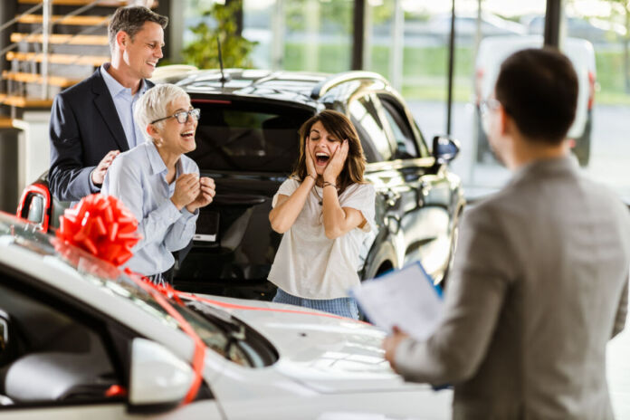 Eine ältere Frau, ein Mann und eine Frau mittleren Alters und ein Autohändler stehen in einem Autohaus. Die jüngere Frau jubelt, weil ihr offensichtlich das weiße Auto mit der roten Schleife herum geschenkt wird. Alle lächeln zufrieden.