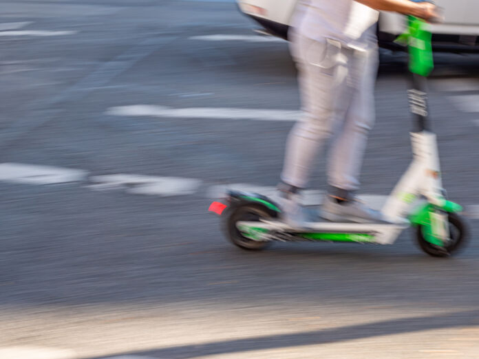 Ein E-Scooter-Fahrer fährt durch die Stadt. Dabei passiert er eine Kreuzung und fährt an Autos und Fahrrädern vorbei. Die Situation im Verkehr kann gefährlich sein.