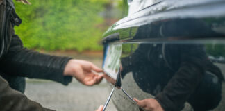 Die Nahaufnahme eines Nummernschildes am Auto und einer Hand, die versucht, dieses mithilfe eines Schraubenziehers zu demontieren oder zu stehlen.