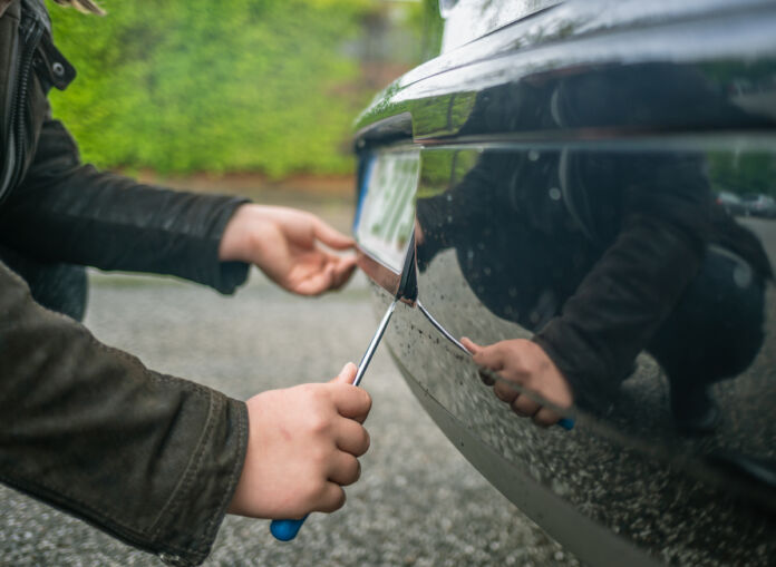 Die Nahaufnahme eines Nummernschildes am Auto und einer Hand, die versucht, dieses mithilfe eines Schraubenziehers zu demontieren oder zu stehlen.