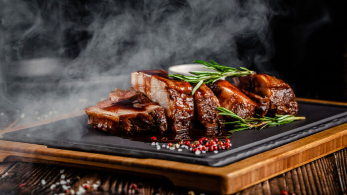 Ein Schiefertafel mit Grillfleisch in einem Grillrestaurant dampft appetitlich. Dazu gibt es Rosmarin als Gewürz, das Fleisch wurde auf dem Stein erhitzt.