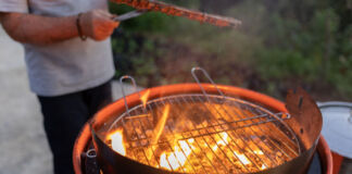 Ein Mann grillt ein Stück Fleisch auf einem Holzkohle-Grill. Flammen wirbeln auf der Kohle unter dem Grillrost. Der Mann hält mit der Zange das Steak fest. Im Hintergrund ist ein Garten.