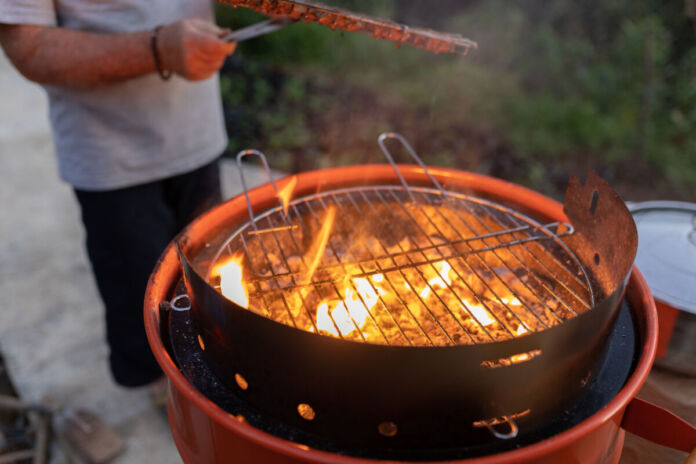 Ein Mann grillt ein Stück Fleisch auf einem Holzkohle-Grill. Flammen wirbeln auf der Kohle unter dem Grillrost. Der Mann hält mit der Zange das Steak fest. Im Hintergrund ist ein Garten.