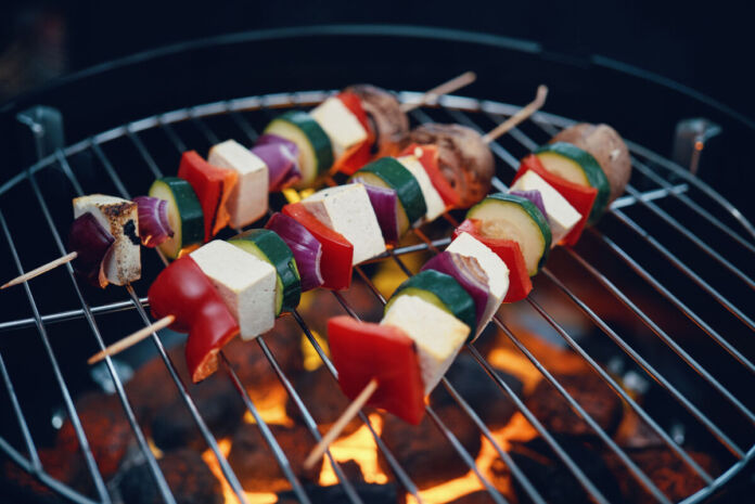 Auf dem Grillrost eines Grills, der mit Kohle befeuert wird, befinden sich drei vegane Spieße. Diese bestehen aus Tofu, Paprika, Zwiebeln, Zucchini und Champignons.