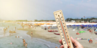 Die Hand einer Frau hält ein Thermometer ins Bild. Im Hintergrund sieht man viele Urlauber am Strand liegen und im Meer schwimmen.