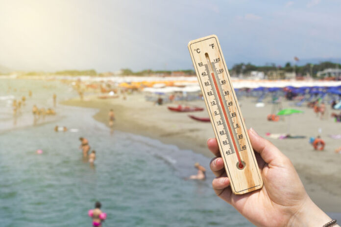 Die Hand einer Frau hält ein Thermometer ins Bild. Im Hintergrund sieht man viele Urlauber am Strand liegen und im Meer schwimmen.