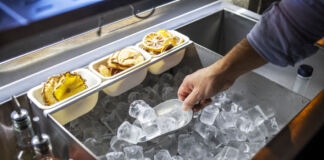 Ein Barkeeper nimmt mit einer Schaufel einige Eiswürfel aus einem großen Becken mit vielen Eiswürfeln. Über dem Eis liegen in hängenden kleineren Behältern getrocknete Zitronenscheiben.