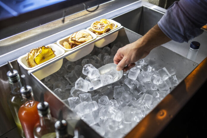 Ein Barkeeper nimmt mit einer Schaufel einige Eiswürfel aus einem großen Becken mit vielen Eiswürfeln. Über dem Eis liegen in hängenden kleineren Behältern getrocknete Zitronenscheiben.