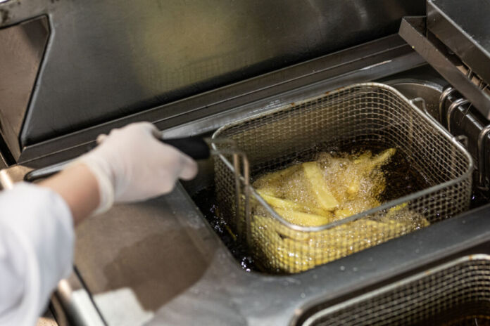 Ein Mitarbeiter oder eine Mitarbeitern hält ein Behälter mit Pommes frites in das Frittieröl in das Becken. Er oder sie trägt Handschuhe.