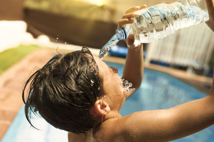 Ein Junge gießt sich Wasser aus einer Flasche über den Kopf, um sich abzukühlen. Es herrscht offensichtlich Hitze im Sommer. Im Hintergrund ist ein Swimmingpool, und die Sonne scheint.