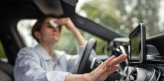 Eine Frau sitzt in einem Auto. Sie ist sichtlich verschwitzt und es ist heiß. Sie wischt sich den Schweiß von der Stirn, denn das Auto hat eine kaputte Klimaanlage.