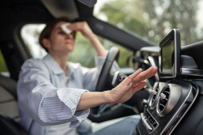 Eine Frau sitzt in einem Auto. Sie ist sichtlich verschwitzt und es ist heiß. Sie wischt sich den Schweiß von der Stirn, denn das Auto hat eine kaputte Klimaanlage.