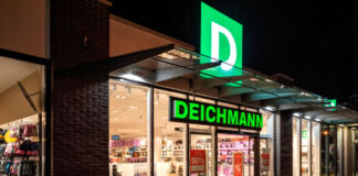 Deichmann bei Nacht. Eine Filiale des Schuhladens in einer Fußgängerzone ist beleuchtet, während es draußen dunkel ist. Man erkennt das Angebot sowie den grünen Schriftzug und das Logo des Konzerns.