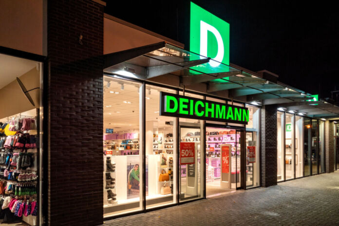 Deichmann bei Nacht. Eine Filiale des Schuhladens in einer Fußgängerzone ist beleuchtet, während es draußen dunkel ist. Man erkennt das Angebot sowie den grünen Schriftzug und das Logo des Konzerns.