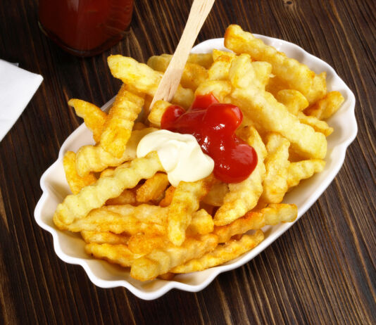 Eine Portion Pommes frites steht auf einem Tisch mit einer Holzplatte. Direkt auf den Pommes frites befinden sich zwei Spritzer Ketchup und Mayonnaise.