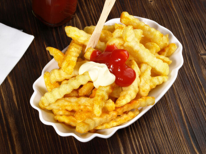 Eine Portion Pommes frites steht auf einem Tisch mit einer Holzplatte. Direkt auf den Pommes frites befinden sich zwei Spritzer Ketchup und Mayonnaise.