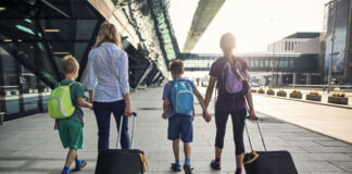 Eine Mutter mit drei Kindern läuft an einem Flughafen entlang. Das Mädchen und die Mutter ziehen einen Koffer hinter sich her. Alle tragen Rucksäcke.