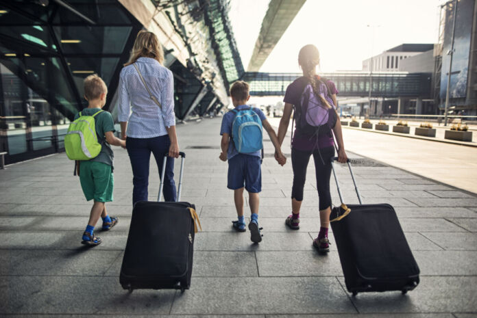 Eine Mutter mit drei Kindern läuft an einem Flughafen entlang. Das Mädchen und die Mutter ziehen einen Koffer hinter sich her. Alle tragen Rucksäcke.