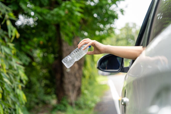 Eine Frauenhand hält eine Plastikflasche in der Hand und ist im Begriff, diese aus dem Fenster zu werfen. Das Auto fährt und bewegt sich auf einer Straße durch die Natur.