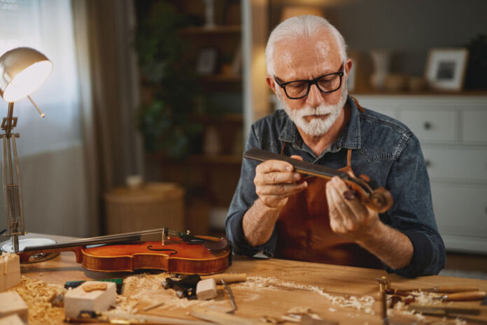 Ein älterer Mann stellt an einem Tisch eine Geige aus Holz her. Man sieht, dass dieser Prozess noch aus reiner Handarbeit besteht.