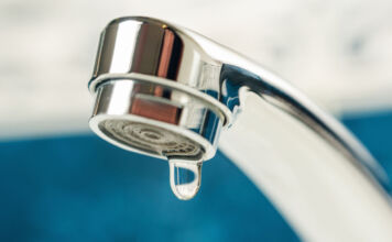 Nahaufnahme eines Wasserhahns mit Wassertropfen. Der Wassertropfen hängt an dem silbernen Kopf des Wasserhahns für Trinkwasser. Womöglich ist der Wasserhahn undicht, weshalb der Tropfen herunterzufallen droht.