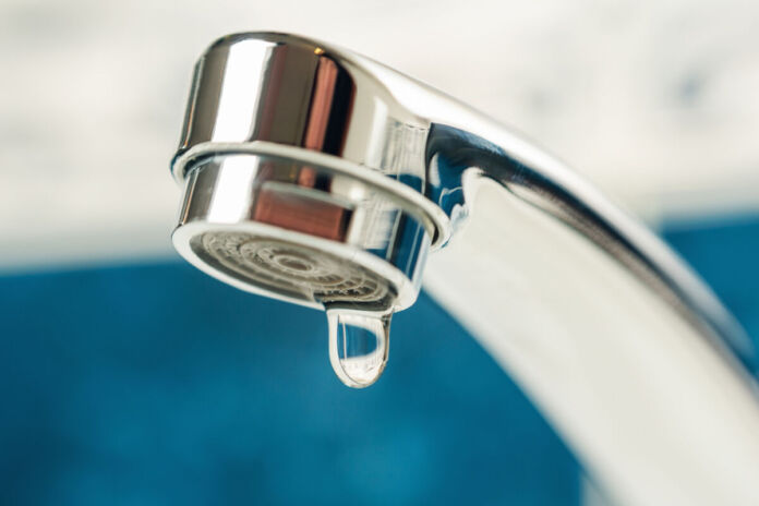 Nahaufnahme eines Wasserhahns mit Wassertropfen. Der Wassertropfen hängt an dem silbernen Kopf des Wasserhahns für Trinkwasser. Womöglich ist der Wasserhahn undicht, weshalb der Tropfen herunterzufallen droht.